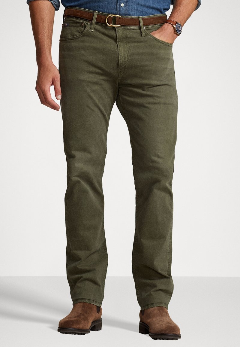 Chinos de color verde oliva, tejido de algodón, diseño de pierna recta, cinco bolsillos, cinturón de cuero marrón con hebilla de metal, llevados con botas de ante marrón.