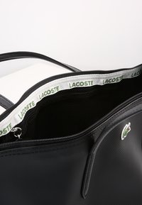 Schwarze Tragetasche mit Reißverschluss, die eine strukturierte Oberfläche, ein Stofffutter und ein grünes Lacoste-Logo aufweist. Innen ist eine Netz-Organisertasche enthalten.