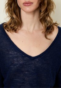 Tamsiai mėlynas, lengvas megztinis su V formos iškirptė ir tekstūruota apdaila. Turi laisvą siluetą, demonstruojantį minkštą, šiek tiek permatomą medžiagą.