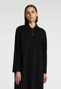 Zwarte lange mouwen polo jurk van zachte stof, met een klassieke kraag en drie ronde knopen aan de voorkant. Eenvoudige en ontspannen pasvorm.