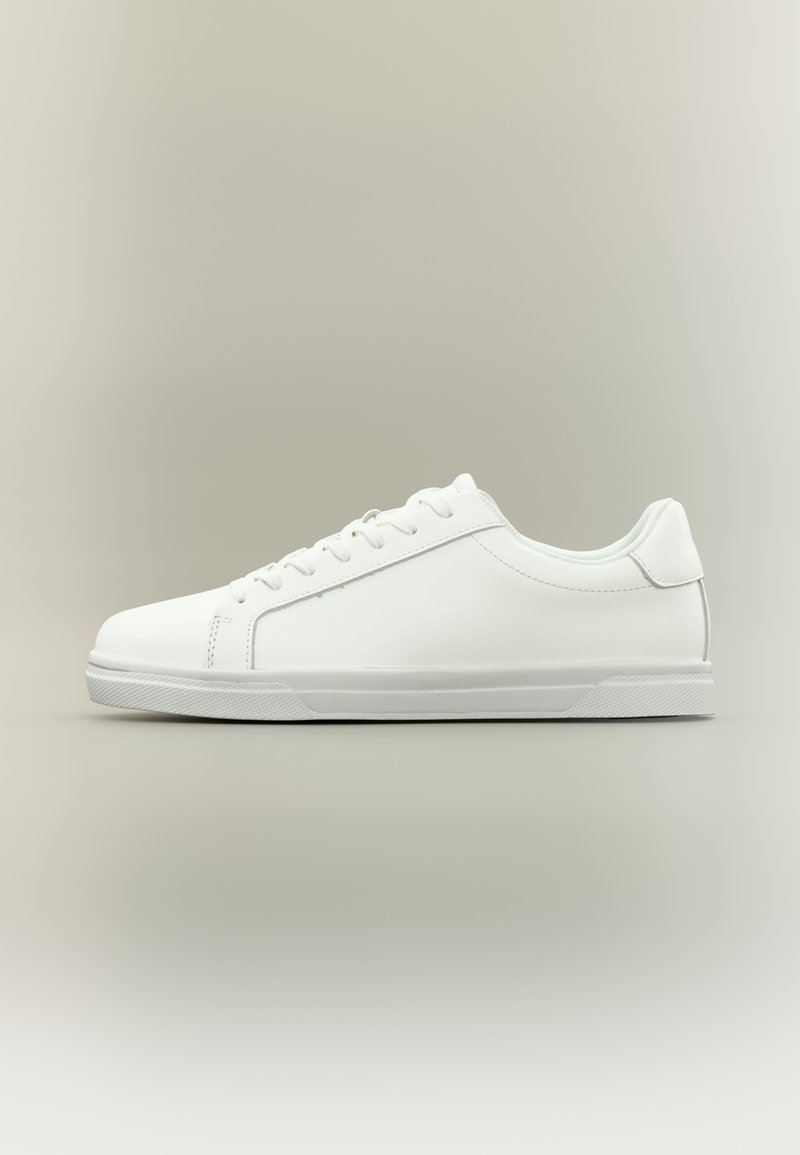 Λευκό αθλητικό παπούτσι χαμηλού τύπου με κορδόνια μπροστά και υφασμένο λάστιχο στη σόλα, σε ουδέτερο απλό φόντο.