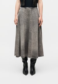 Γυναίκα που φοράει μακριά φούστα σε γκρι απόχρωση με εφέ οξειδωμένης όξινης πλύσης, με ένα μικρό μεταλλικό λογότυπο στη μέση και μαύρες μπότες με μυτερή μύτη.