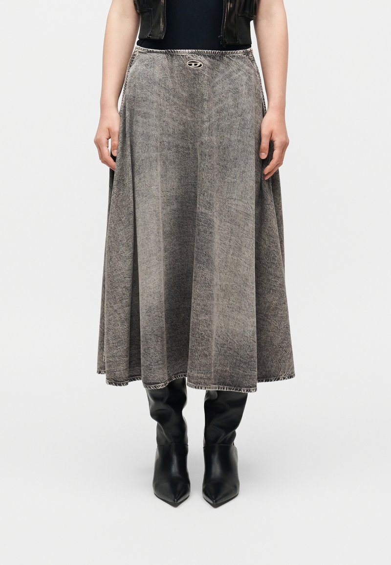 Γυναίκα που φοράει μακριά φούστα σε γκρι απόχρωση με εφέ οξειδωμένης όξινης πλύσης, με ένα μικρό μεταλλικό λογότυπο στη μέση και μαύρες μπότες με μυτερή μύτη.