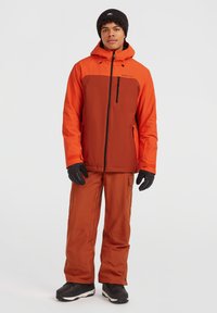 Orange og brun skijakke med en lynlåslomme, parret med matchende brune bukser. Model iført sorte handsker og støvler.