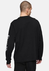 Camiseta negra de manga larga hecha de algodón, con cuello redondo y mangas hasta el codo. Texto "Sportlife" en blanco en la manga izquierda.
