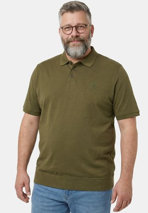 Średni mężczyzna z brodą i okularami, ubrany w oliwkową koszulkę polo i jasnoniebieskie dżinsy, stojący na jasnym tle.
