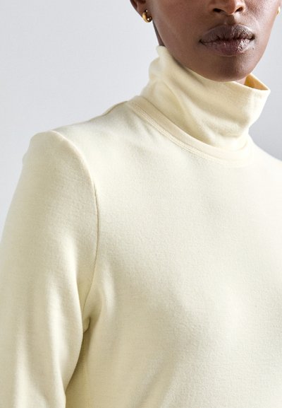 Gaišdzeltena turtlenecka džemperis, kas izgatavots no mīkstas, teksturētas auduma ar garām piedurknēm un pieguļošu siluetu. Šuves detalizējums gar apkakli.