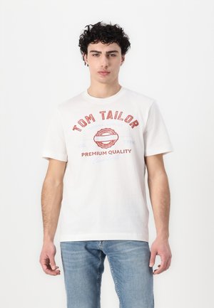 Bijela pamukasta majica s kratkim rukavima, s crvenim natpisom "TOM TAILOR" i dekorativnim grafičkim elementima. Glatka tekstura, klasičan kroj.