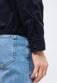 Inimese käe lähivõte, kes kannab musta nööbitud jopeserva ning näha on hele sinist teksapükste taskut.