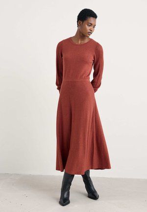Rust-colored long-sleeve dress with a fitted bodice and flared skirt. Knit texture with ribbed detailing on the sleeves. Worn with black knee-high boots.