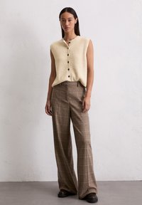 Cremfarbener, ärmelloser Strickoberteil mit Knopfverschluss, kombiniert mit hoch taillierten, braun karierten, weit geschnittenen Hosen. Schwarze Schuhe runden den Look ab.