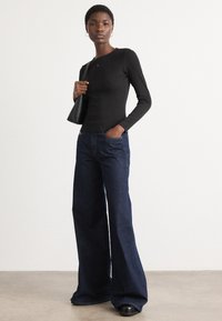 Black long-sleeve top with fitted design, paired with flared dark blue jeans. Model carries a small black bag; standing on a light surface.