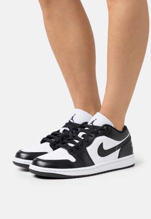 Zapatillas bajas en blanco y negro que combinan materiales de cuero y sintético, con una caja de puntera perforada y los icónicos logos de Nike y Jumpman.