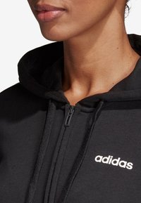 Sweat à capuche zippé noir en tissu doux, doté d'une capuche à cordon de serrage et du logo blanc adidas sur le côté gauche. Gros plan sur l'encolure.