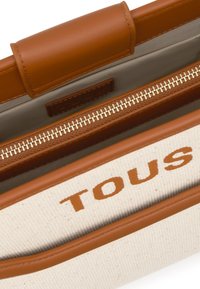 Bolso de lona con detalles en cuero marrón, con cierre de cremallera y un interior claro. "TOUS" está impreso en naranja en el lateral.