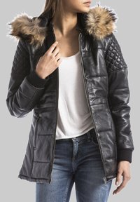 Giubbotto di pelle nera con maniche trapuntate, cappuccio foderato in pelliccia, chiusura con zip e polsini a costine, abbinato a una maglietta bianca e jeans blu.