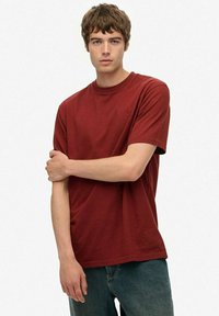 Ung mand med kort brunt hår iført en ensfarvet rød T-shirt og blå jeans, stående med den ene arm på tværs af kroppen.