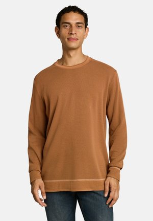 Homme debout portant une chemise marron à manches longues texturée et un jean bleu foncé sur un fond clair uni.
