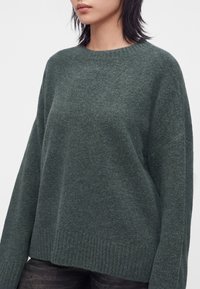 Person iført en mørkegrøn, langærmet striktrøje med rund halsudskæring og ribkant mod en ensartet lys baggrund.