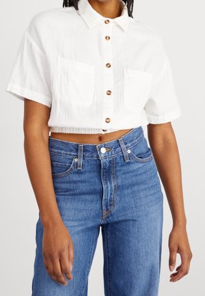 Kvinde iført en hvid kortærmet skjorte med knapper og højtaljede blå jeans, stående foran en ensfarvet baggrund.