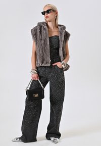 Gilet in pelliccia sintetica grigia sopra una maglia nera a fantasia, abbinato a pantaloni in denim neri decorati con borchie argentate e scarpe argentate. Borsa nera inclusa.