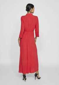 Raudona maksi suknelė su ilgomis rankovėmis, įtemta liemeniu ir klostuota nugara. Dėvima su juodais aukštakulniais. Lygios medžiagos tekstūra.