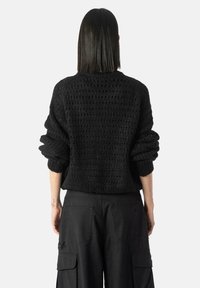 Persoană cu păr drept negru, îmbrăcată într-un pulover texturat negru și pantaloni cargo negri cu mânecile suflecate, întoarsă cu spatele.