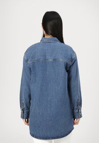 Dolga denim srajca v srednji modri barvi, ki ima koničast ovratnik, gumbaste manšete in zaobljen rob. Teksturirana tkanina z vidnimi šivi.