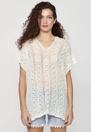 Poncho blanc en tricot avec un col en V et des manches courtes, comportant un motif ajouré détaillé et des rayures horizontales, tombant de manière fluide.