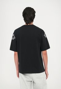 Μαύρη βαμβακερή μπλούζα με κοντά μανίκια, με γραφικό σχέδιο στο επάνω αριστερό μανίκι και άνετη εφαρμογή.