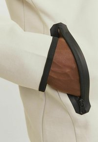 Veste beige clair avec une poche zippée noire dotée d'un bord en caoutchouc texturé, révélant une main à la peau brune à l'intérieur de la poche.