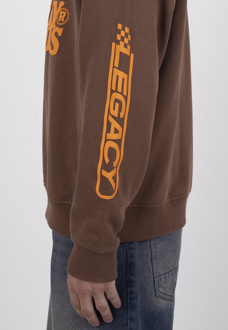 Manche de sweat-shirt marron avec texte orange "LEGACY" et motif à carreaux porté avec un jean en denim bleu sur le bras d'une personne.