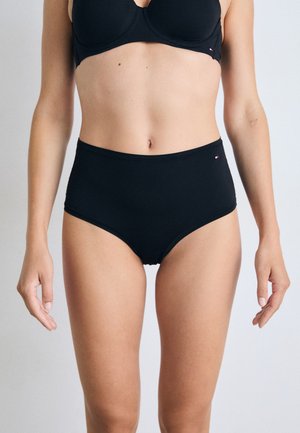 Femme portant un ensemble noir assorti de soutien-gorge et de culotte taille haute avec un petit logo sur la hanche, se tenant devant un fond clair.