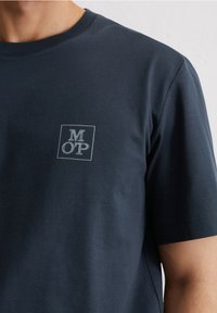 Marineblaue T-Shirt aus weichem Baumwollstoff, mit einem quadratischen Logo, das "M OP" in weißer Stickerei auf der oberen linken Brust zeigt. Kurze Ärmel.