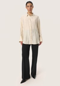 Cămașă cu o textură moale, de culoare crem deschis, cu guler și manșete lungi, cu o croială puțin relaxată. Asortată cu pantaloni largi, cu croială adaptată, de culoare neagră și pantofi cu vârful ascuțit.