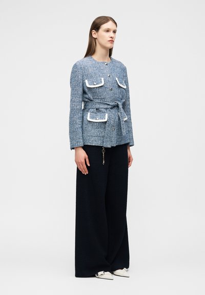 Γυναίκα που φοράει μπλε υφασμάτινο σακάκι με ζώνη και λευκές λεπτομέρειες, και μαύρο παντελόνι με φαρδιά μπατζάκια, στέκεται μπροστά σε απλό ανοιχτό φόντο.