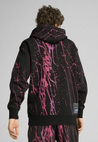 Sudadera negra con un diseño de salpicaduras en púrpura, cuenta con un bolsillo tipo canguro, puños y dobladillo acanalados. Detalles de la etiqueta visibles en el lado inferior.