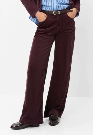 Personne portant un pantalon large en velours côtelé bordeaux foncé, une ceinture noire, une chemise rayée bleue et des mocassins noirs, les mains dans les poches.