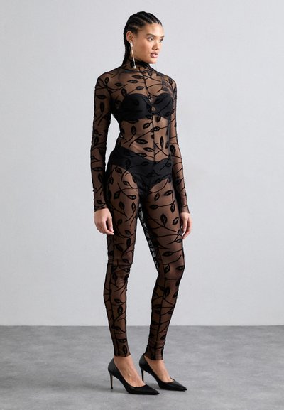 Bodysuit noir transparent avec motif de feuilles, manches longues, col montant et sous-vêtements noirs, associé à des chaussures à talons noires.
