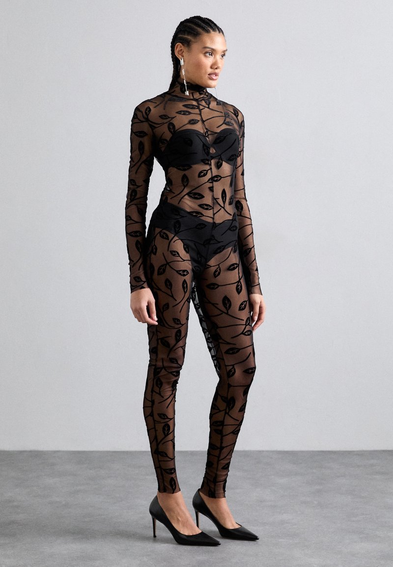 Sheer black bodysuit with leaf pattern, long sleeves, high neck, and black undergarments, paired with black heeled shoes.