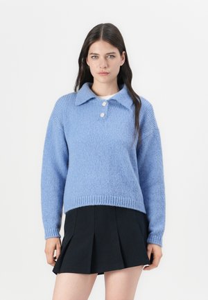 Jasnoniebieski dzianinowy sweter z kołnierzykiem, trzema guzikami i ściągaczami przy mankietach. Noszony z czarną plisowaną spódnicą.