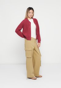 Κόκκινη πλεκτή ζακέτα με στρογγυλή λαιμόκοψη, κλείσιμο με κουμπιά και ριμπ μανσέτες, συνδυασμένη με μπεζ cargo παντελόνια που διαθέτουν μεγάλες πλαϊνές τσέπες.