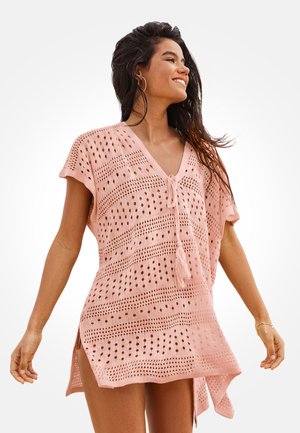 Kobieta uśmiechająca się, ubrana w luźną, różową, dzianą plażową narzutkę z dekoltem w serek i troczkami z frędzlami, stojąca z ramionami swobodnie opuszczonymi na tle białego tła.