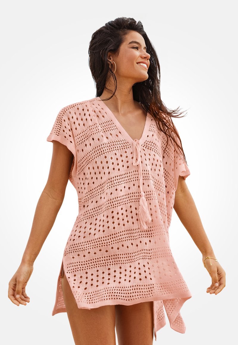 Mujer sonriente, vistiendo una cubierta de playa holgada de punto rosa con escote en V y cordones con borlas, de pie con los brazos relajados contra un fondo blanco.