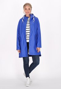 Kék vízálló dzseki, húzózsinóros kapucnival, cipzáras elejével, patentgombokkal és oldalsó zsebekkel. Csíkos póló és farmernadrág fölött viselve.