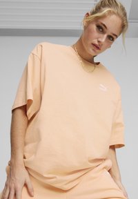 Lys ferskenfarvet bomulds-T-shirt med afslappet pasform, korte ærmer og et lille hvidt logo på brystet. Kombineret med matchende ferskenfarvede bukser.