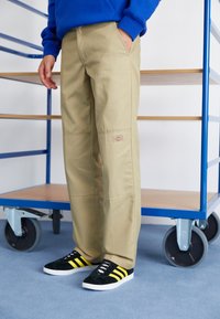 Pantalones de pierna ancha en color beige con dos bolsillos laterales, un parche de logo cosido, combinados con zapatillas negras que tienen rayas amarillas sobre un fondo azul.