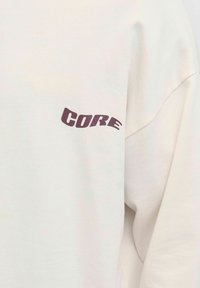 Cremefarvet sweatshirt med løs pasform, med ordet "CORE" i mørk burgundy på den venstre ærme, lavet af blødt bommateriale.