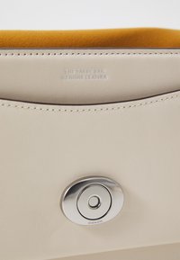 Cremfarbene Ledertasche mit geliner Wildlederauskleidung. Verfügt über einen runden silbernen Verschluss und eingeprägten Text mit der Aufschrift "DIE TABBY-TASCHE ECHTES LEDER".