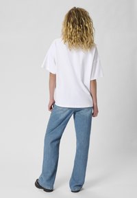 White oversized cotton t-shirt with short sleeves, paired with light blue wide-leg denim jeans and black footwear, viewed from the back.